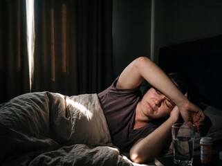 Fototapeta premium A close-up of a woman suffering from a migraine lying in bed in a completely darkened room where a single blade of light cuts through the dimness and the woman covers her eyes with the back of her han