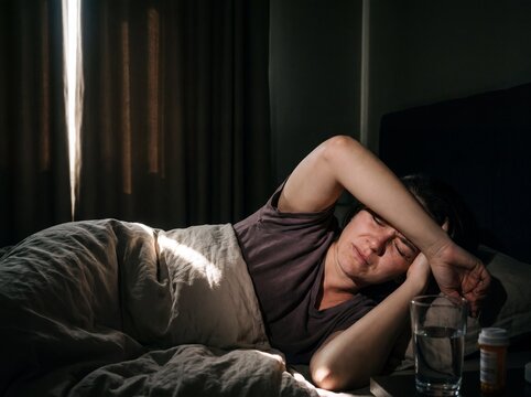 A close-up of a woman suffering from a migraine lying in bed in a completely darkened room where a single blade of light cuts through the dimness and the woman covers her eyes with the back of her han