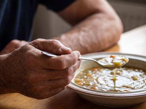 A macro detail of a hand affected by Parkinson's disease trying to hold steady a spoon full of soup, the shaky camera movement following the rhythmic and uncontrollable tremor of the hand, highlightin