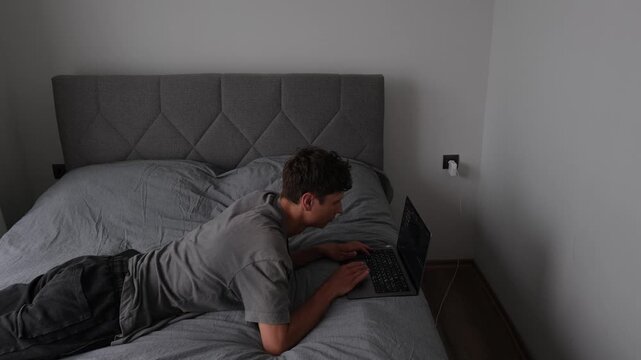 Young freelancer working from bed with laptop in small apartment, realistic remote work lifestyle in imperfect home office conditions and limited living space
