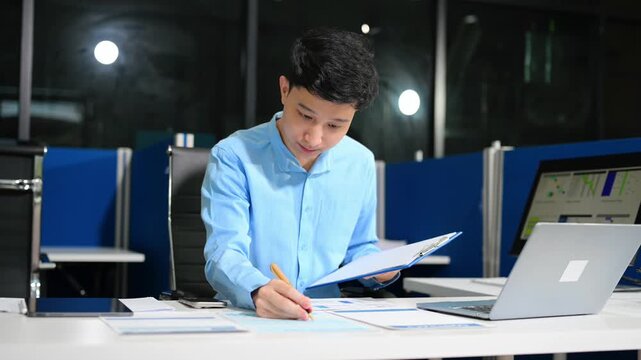 Young Asian businessman using a smartphone app for digital verification while working on a laptop late