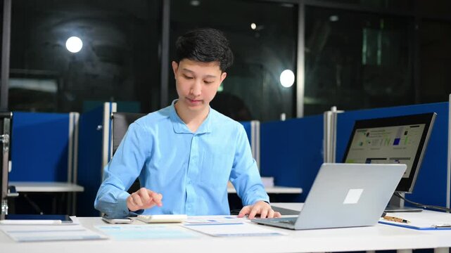 Asian businessman checking data report on a laptop and writing notes on a clipboard in the office.