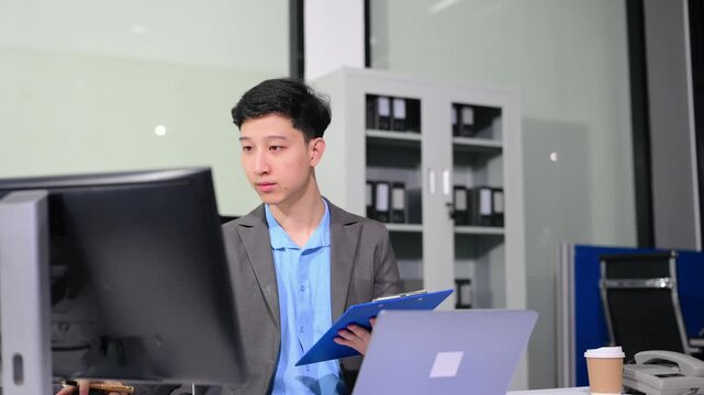 Asian businessman checking data report on a laptop and writing notes on a clipboard in the office.