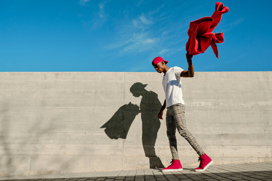 Young man holding red jacket and walking against concrete wall