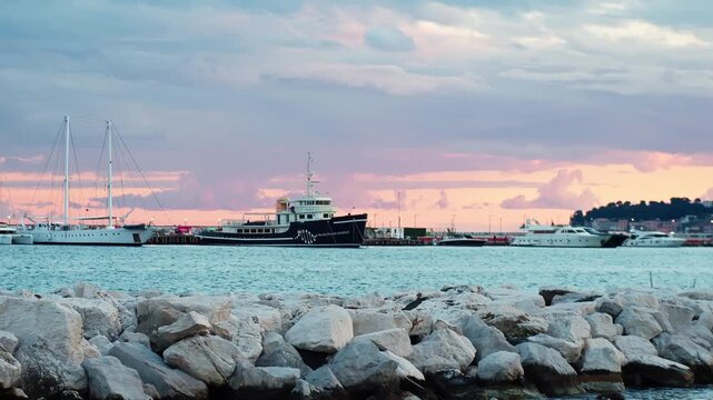Black vintage-style yacht and sailboats moored in Naples at sunset, framed by white shoreline rocks, calm bay water, and a pastel evening sky.