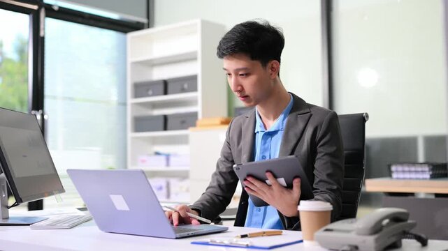 Asian businessman checking data report on a laptop and writing notes on a clipboard in the office.