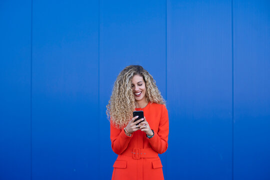 Portrait of young woman wearing red dress using cell phone in front of blue background
