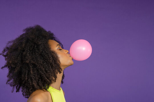 Young woman with eyes closed blowing pink bubble gum against purple background