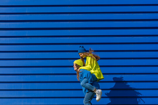 Teenage girl giving her friend a piggyback ride in front of blue background