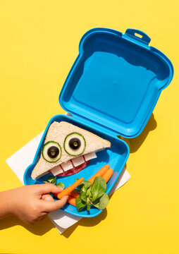Hand of baby girl picking up baby carrot from lunch box with funny looking sandwich with anthropomorphic face