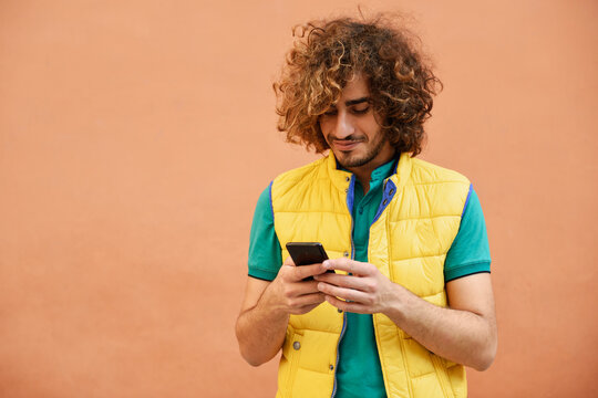 Smiling young man with curly hair wearing yellow waistcoat looking at cell phone