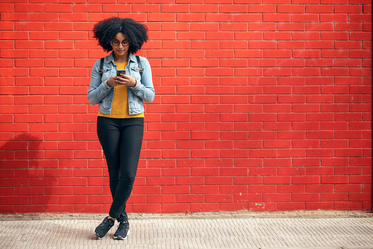 Young woman using smart phone while standing in front of red brick wall