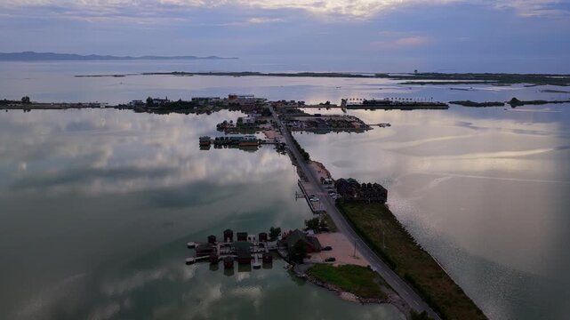 Aerial video over  a straight roadway network connecting modern resort complexes, parking areas, and stilt houses across the mirrored wetlands of Patok Lagoon, Albania.