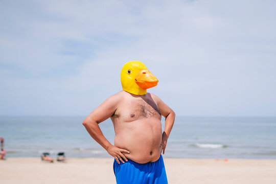 Shirtless obese man with hand on hips wearing duck mask standing at beach