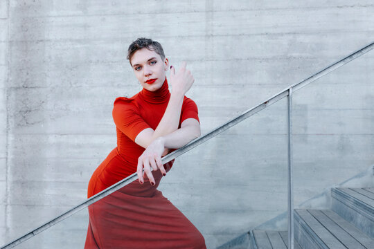 Confident trans man wearing red dress standing on steps against wall