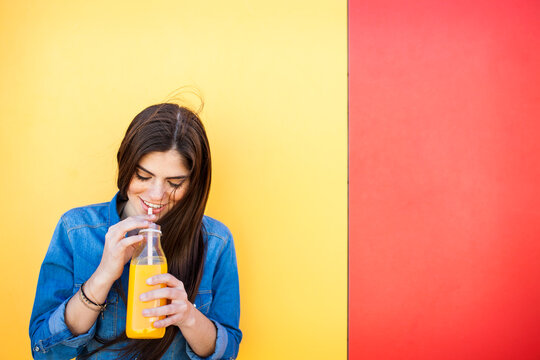 Smiling young woman in front of colourful wall drinking orange juice
