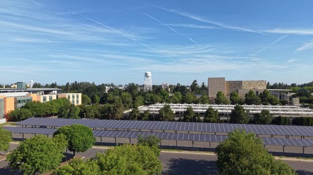 Rising drone view showcasing the UC Davis campus in California, featuring buildings, green trees, and large parking lots covered with solar panels under a bright blue sky