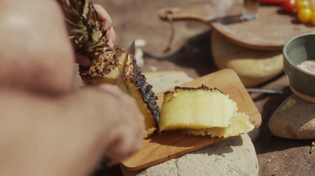 Person slicing grilled pineapple on a wooden cutting board outdoors
