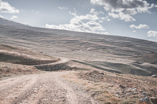 A dusty road on the Pamir Highway winds through mountain hills and slopes among rocks against the backdrop of the Tien Shan mountain peaks