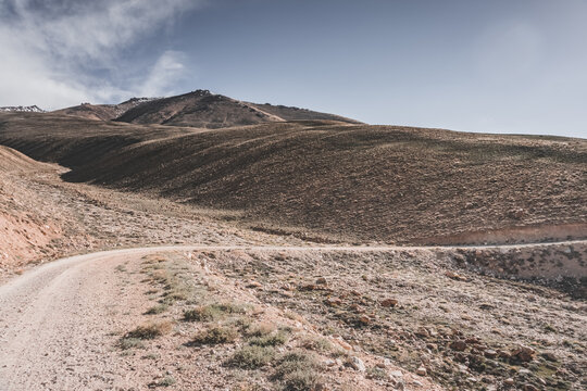 A dusty road on the Pamir Highway winds through mountain hills and slopes among rocks against the backdrop of the Tien Shan mountain peaks