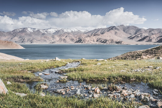 A mountain stream flows down a slope among green grass in sunny summer weather, flowing down to Lake Yashilkul, against the backdrop of a mountain range. Landscape background