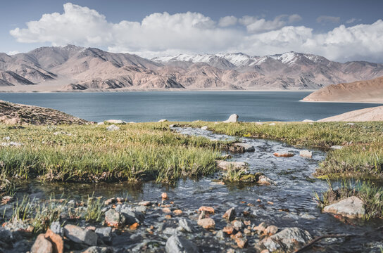 A mountain stream flows down a slope among green grass in sunny summer weather, flowing down to Lake Yashilkul, against the backdrop of a mountain range. Landscape background
