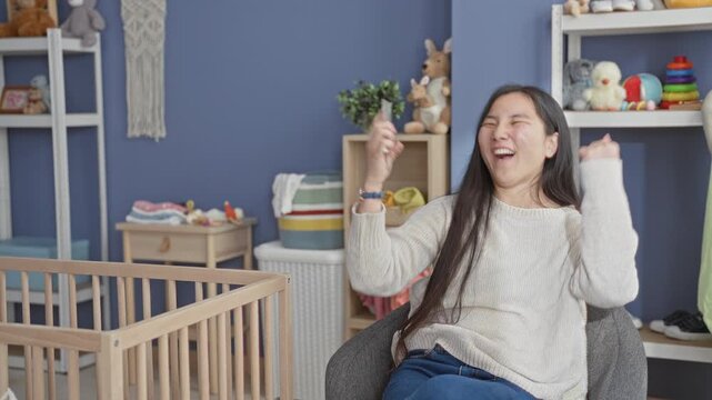 Young chinese woman holding smartphone, smiling and raising hand beside wooden baby crib, toys and storage shelves in a studio playroom; joy playful bonding.