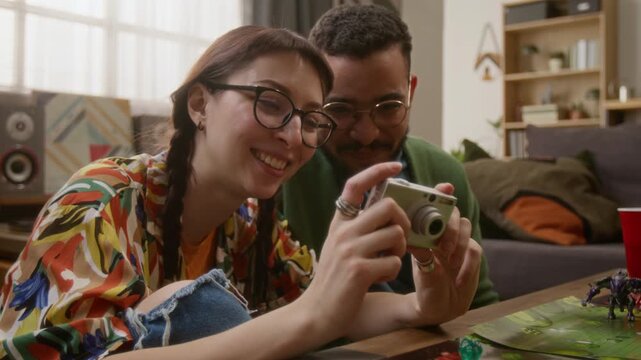 Low angle close up shot of two friends in glasses looking through photos on camera during get-together, playing tabletop games in cozy living room