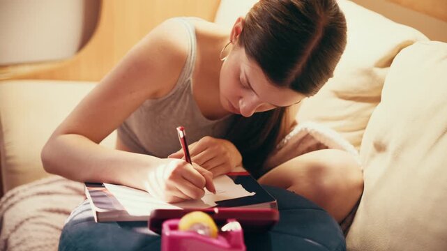Close up of a focused high school student creating secret notes and crib sheets from her smartphone to cheat on a future test, using scissors and tape on a sofa.
