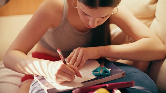 Deeply focused and worried high school girl making secret notes and tiny cheat sheets from her mobile phone, preparing for a test while sitting on a couch under warm indoor lighting.