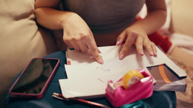Close up of a focused student hand using adhesive tape and scissors on a small piece of paper, finishing a secret crib sheet and notes for a test while sitting on a sofa in a living room.