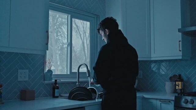 Man standing at kitchen sink washing dishes while snow falls outside the window.