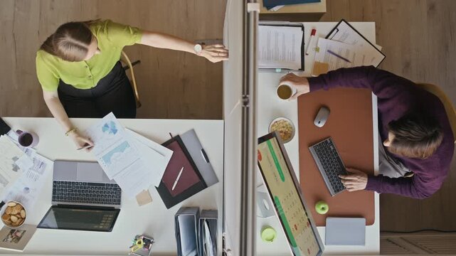 Overhead shot of office coworkers reaching hands across glass divider while working at separate desks in modern workspace