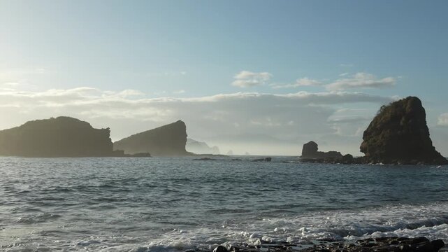Misty tropical seascape featuring large coastal rocks and breaking waves under a bright morning sky at Papuma beach, East Java, Indonesia.