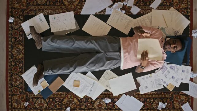 Overhead office scene with exhausted African American male employee lying on office desk surrounded by paperwork after stressful workday