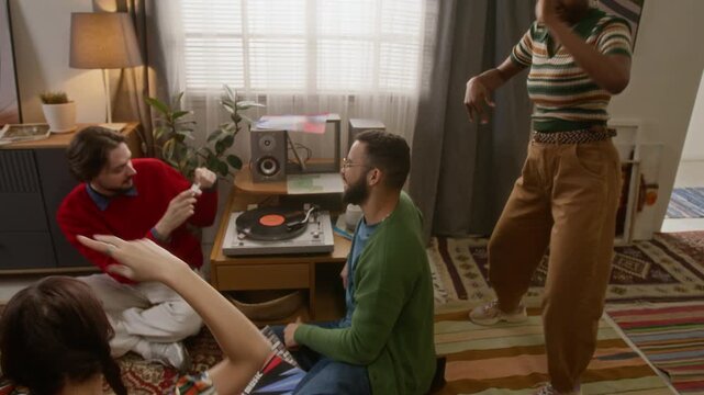 High angle tilt up shot of three friends dancing to vynyl record music while young man filming them with retro camera during cozy party at home