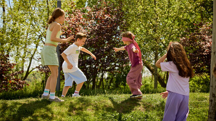 Plakat Children playing blindfold game outdoors in sunny park with happy summer teamwork. Concept of active childhood, outdoor education, summer camp games, friendship, trust and social development.
