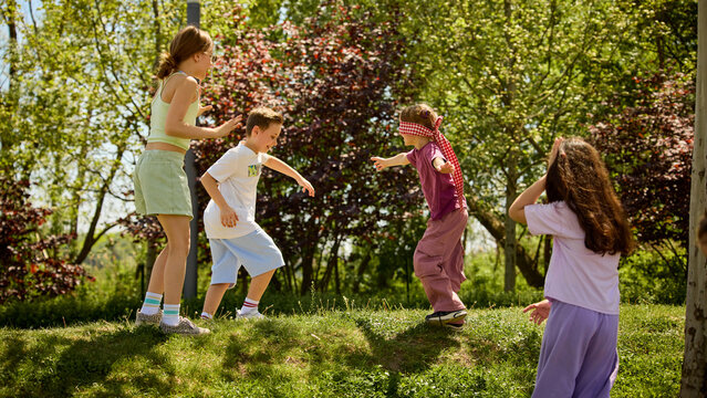 Children playing blindfold game outdoors in sunny park with happy summer teamwork. Concept of active childhood, outdoor education, summer camp games, friendship, trust and social development.