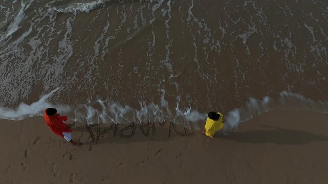 Cinematic high-angle aerial view of the word "Dwarka" written in English on the sea beach sand as ocean waves approach.