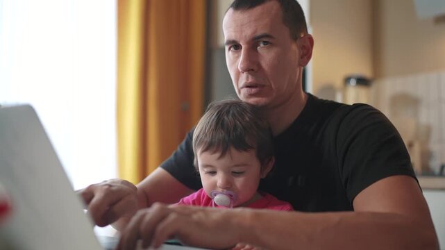 father working from home a remotely with fun baby daughter in his arms. pandemic remote work business concept. father tries to work at home in kitchen, baby children interfere sitting on their hands