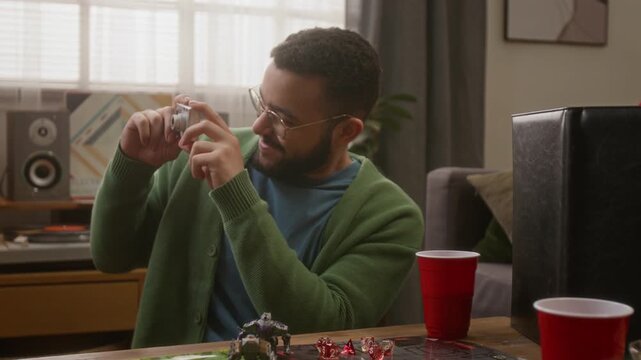 Biracial bearded man in glasses taking pictures of his friends with retro camera while enjoying board game get-together in cozy living room