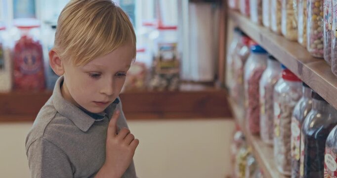 Kid thinking, choice and shopping in a candy store with sweets, confectionery or boy in a retail supermarket, shop and sale of candies. Jar, shelf and child or toddler choosing chocolate product