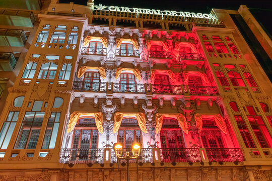 Night view of the illuminated facade of Caja Rural de Aragon building in Zaragoza. The historic architecture is lit with festive red and white lights. Zaragoza, Spain - November 29, 2021
