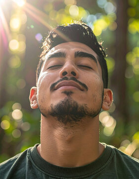 Young man breathing fresh forest air with eyes closed, warm sunlight filtering through trees, peaceful nature therapy concept, environmental health and wellness goals. Close-up.