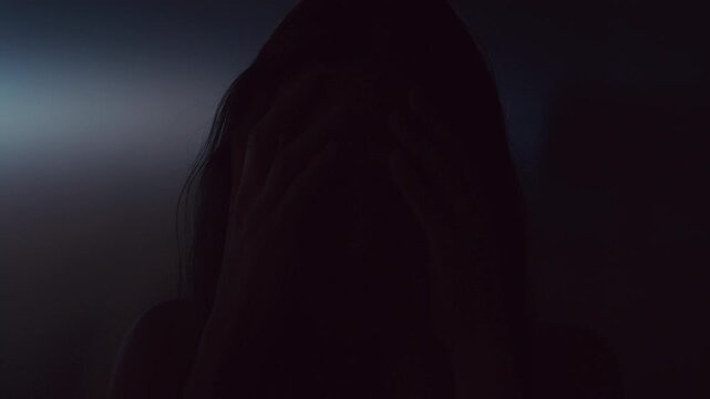 Woman clutching head with both hands and crying out during severe anxiety or panic attack in dark studio with multicolored neon illumination