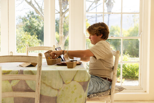 12 year old boy eating a box lunch in his kitchen while using tablet computer