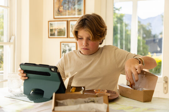 12 year old boy eating a box lunch in his kitchen while using tablet computer