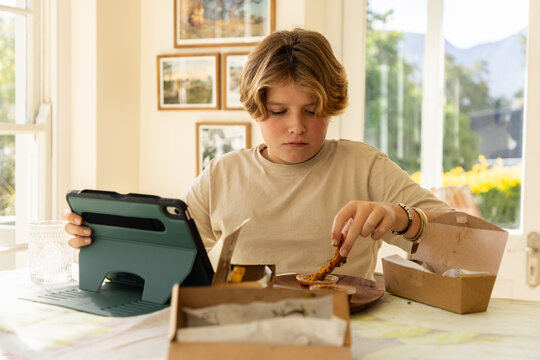 12 year old boy eating a box lunch in his kitchen while using tablet computer