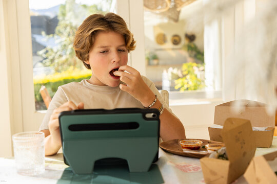 12 year old boy eating a box lunch in his kitchen while using tablet computer
