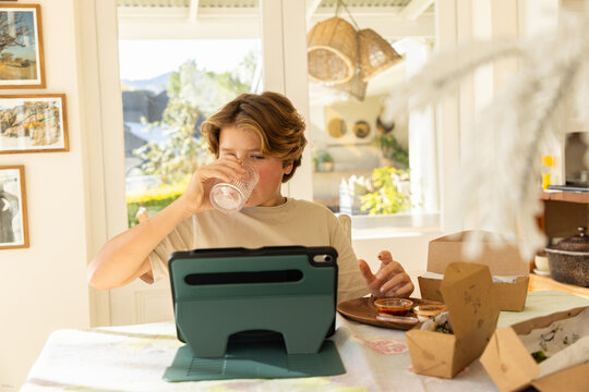 12 year old boy eating a box lunch in his kitchen while using tablet computer
